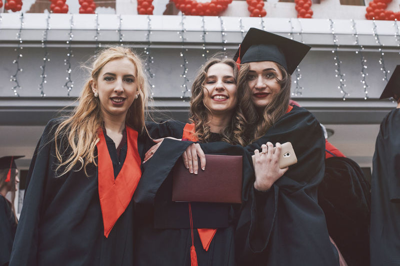 Low angle portrait of happy female students wearing graduation gowns against building