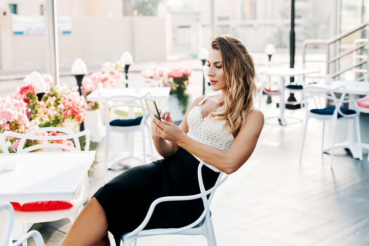 A young stylish woman sits in a street cafe and uses her phone to exchange messages 