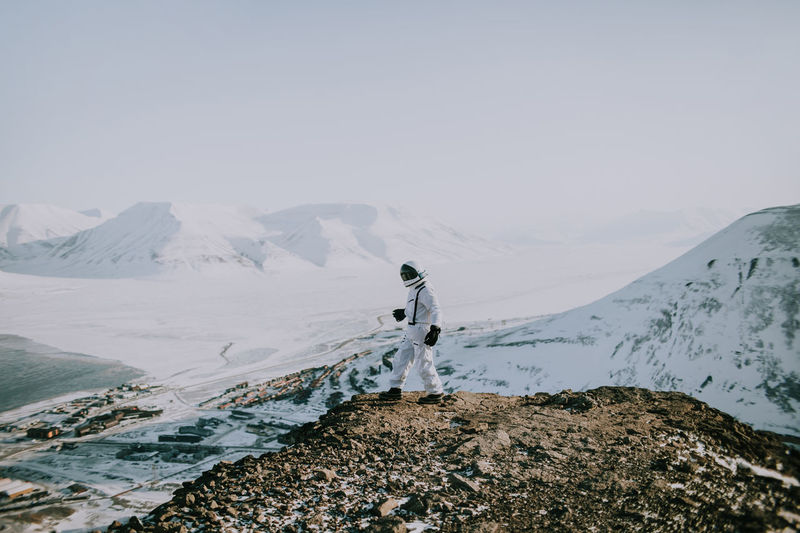 Unrecognizable cosmonaut wearing white spacesuit standing on edge of rocky mountain in winter and admiring amazing landscape in svalbard