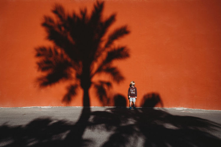 Boy standing against a red wall with the shadow of a palm tree