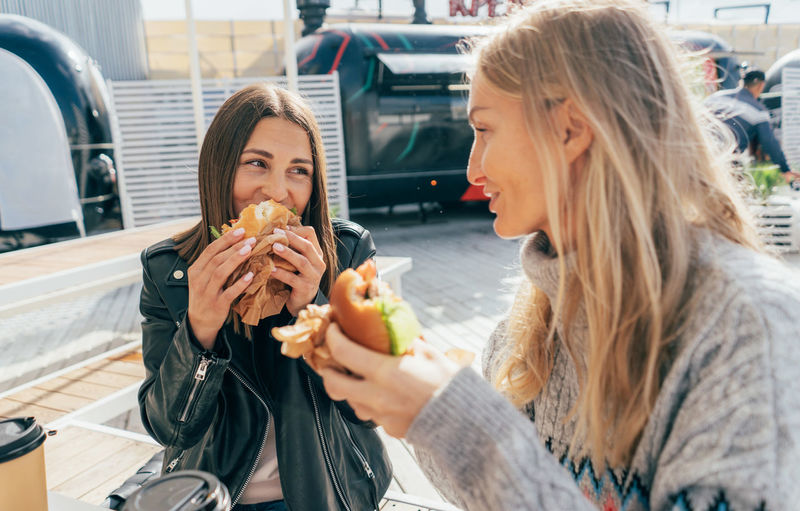 Two attractive european women eat street fast food while sitting at a table outside.