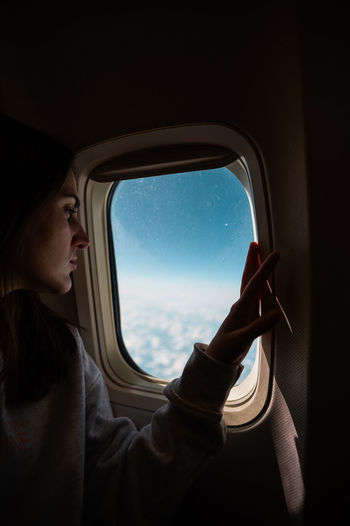 Thoughtful woman admiring cloudy blue sky through window of dark plane cabin