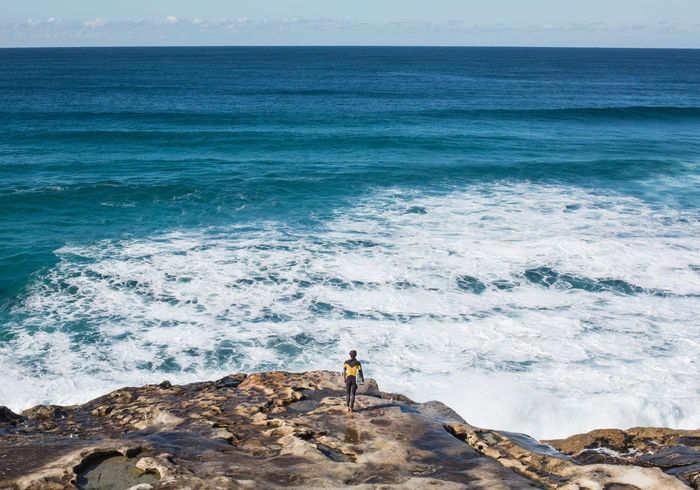 Rear view of male surfer standing on rock overlooking sea