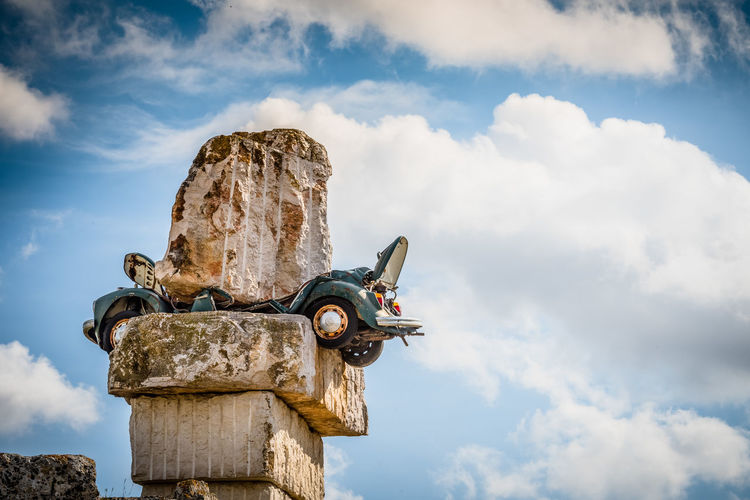 Low angle view of car crushed under rocks against cloudy sky