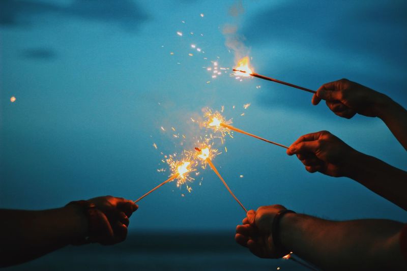 Cropped hands holding lit sparkler against sky