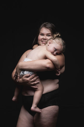 Young plus size woman in underwear holding cute little child in arms and looking at camera with happy smile while standing against black background