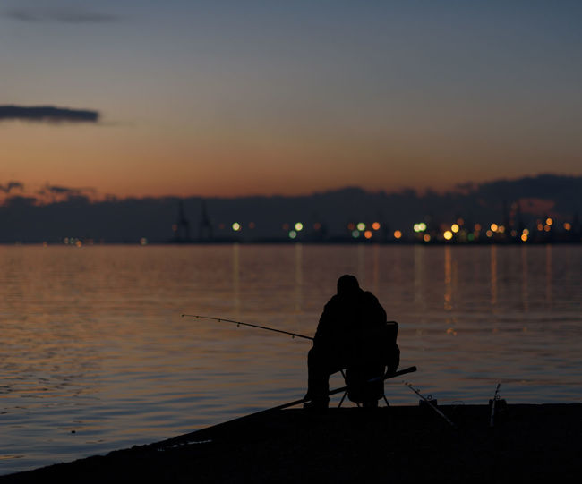 Rear view of silhouette man fishing in sea against sunset sky 