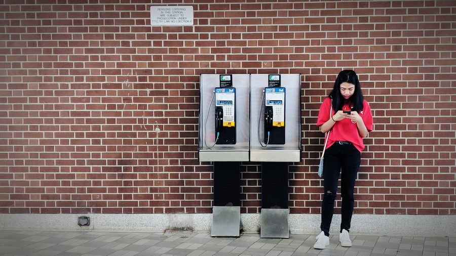Full length of young woman using phone by telephone booth against brick wall