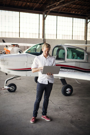 Man using laptop computer while standing by propeller airplane at hangar