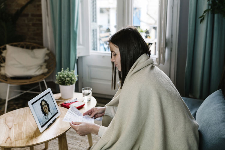 Sick woman wrapped in blanket talking during telemedicine through digital tablet at home
