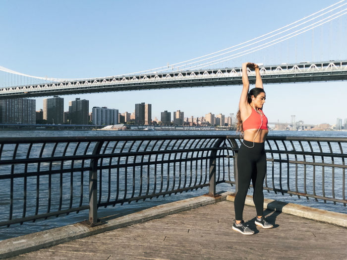 Young woman with arms raised exercising on footpath against bridge in city