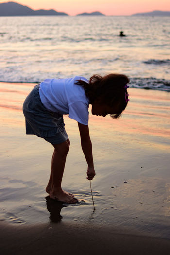 Side view full length of girl bending on shore at beach during sunset