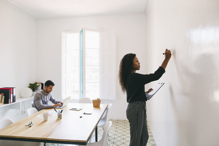 Young businesswoman preparing meeting, writing on wall, colleague working in backgound