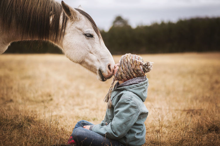 Young girl kisses white horse on nose while sitting on ground