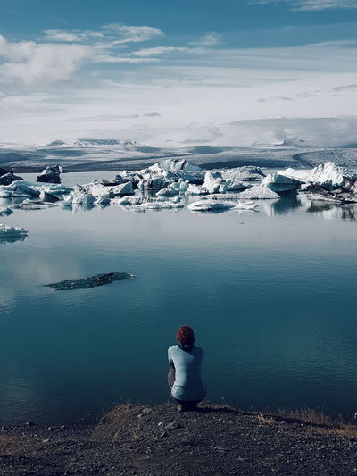 Rear view of woman sitting on by lake during winter