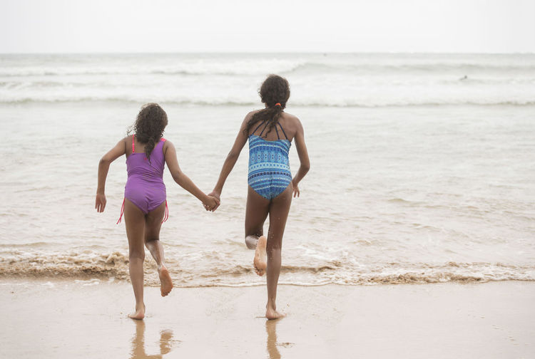 Full length rear view of sisters holding hands while running towards sea on shore at beach