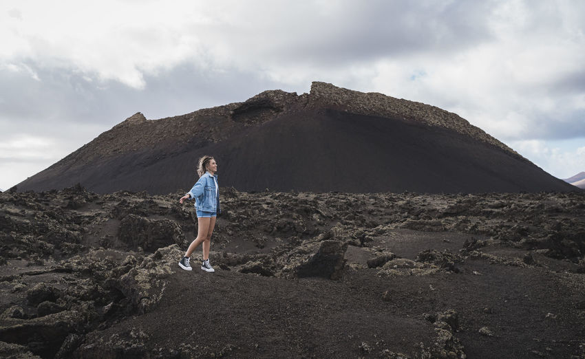 Female tourist looking away while standing with arms outstretched on mountain at volcano el cuervo, lanzarote, spain