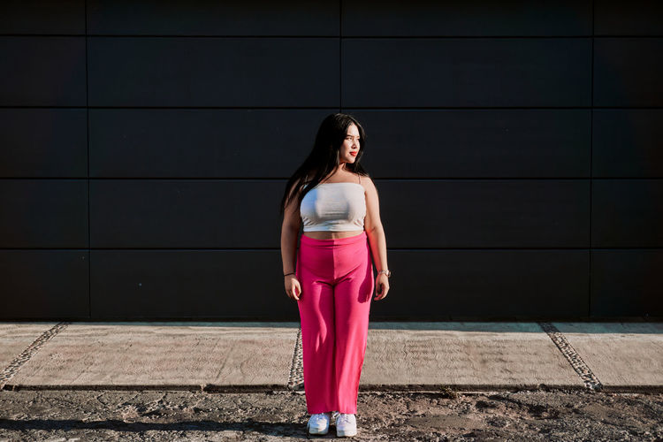 Full body chubby young woman in stylish pink pants and white top looking away while standing against black wall of contemporary building on sunlit city street