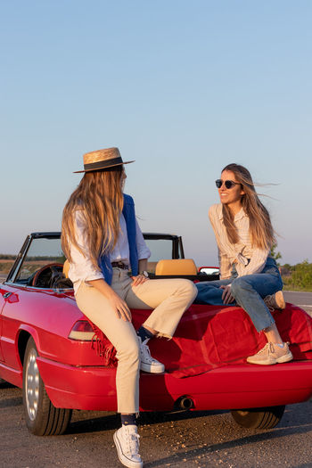 Vertical photo of two teenagers sitting on a convertible red vintage car enjoying their girls trip