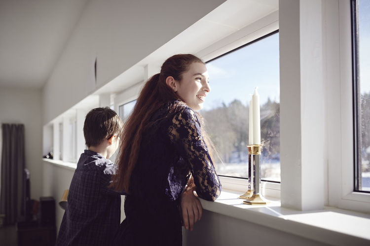 Smiling girl standing with brother while looking through window at home