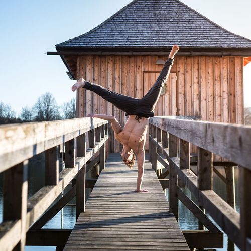 Shirtless man practicing handstand on pier over lake