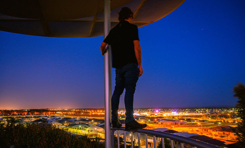 Man standing by illuminated city against clear blue sky at night