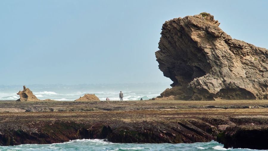 Figure dwarfed by rocks on sea shore against clear sky