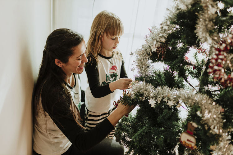 Mother with daughter decorating christmas tree against wall at home