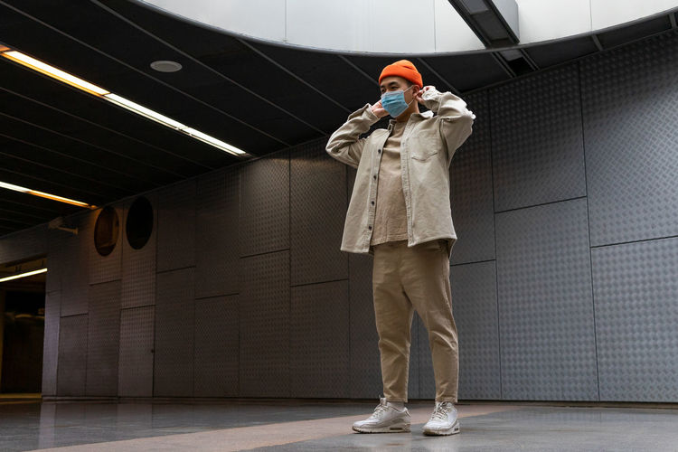 Full body focused stylist asian male wearing casual outfit and face mask standing on concrete building hallway looking away