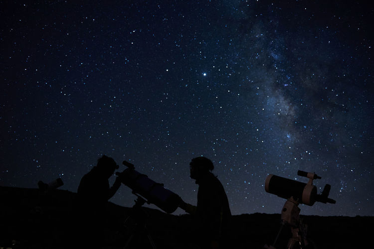 Silhouettes of anonymous scientists using telescopes while exploring night starry sky with milky way in darkness