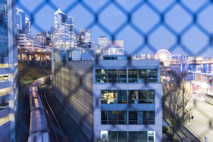 Illuminated buildings in city seen through chainlink fence at dusk