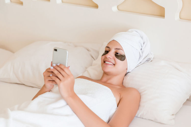 Smiling young woman with under eye patches using mobile phone on bed at spa