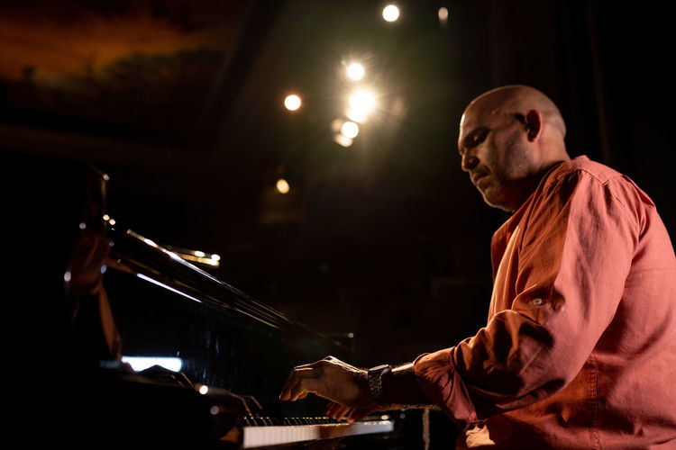 From below side view of bald male musician playing piano during performance on obscure stage in theater