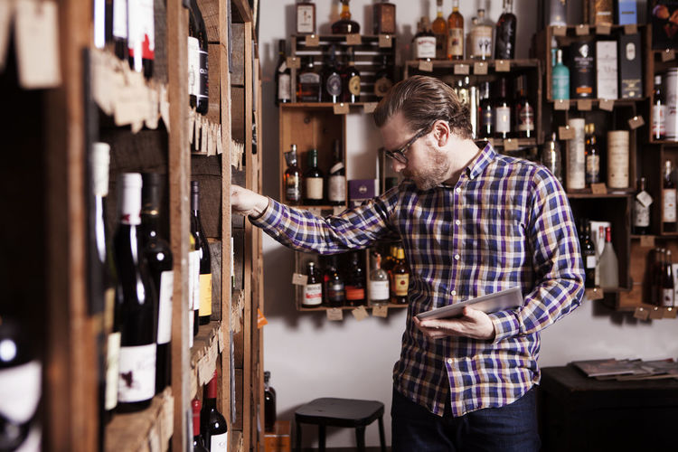 Male small business owner examining wine bottle while holding tablet computer in shop