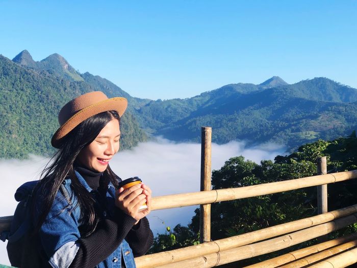 Smiling young woman drinking coffee while standing by railing against mountain