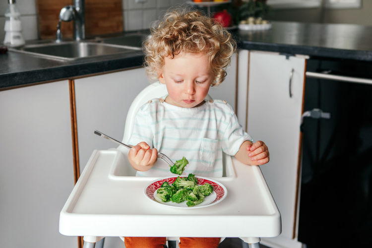 Kid boy sitting in high chair eating broccoli with fork. healthy eating for kids children. 