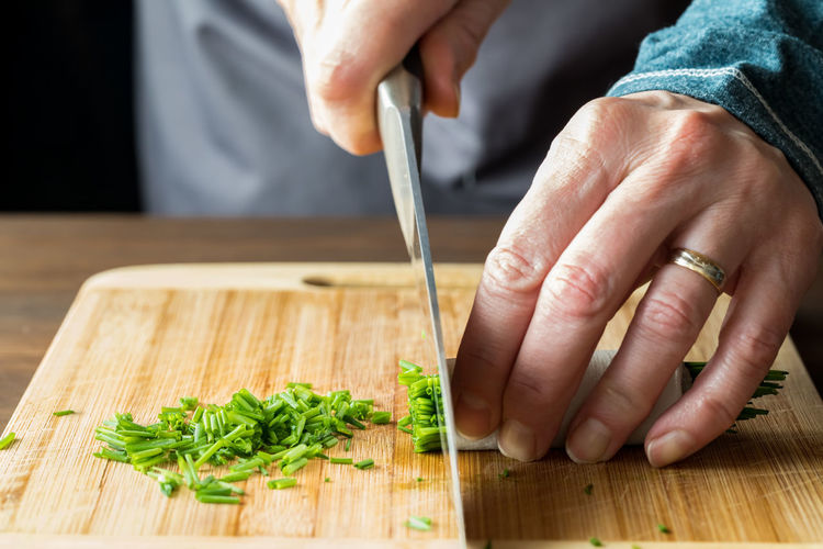 A chef using a sharp knife to chop fresh chives, on a wooden cutting board.