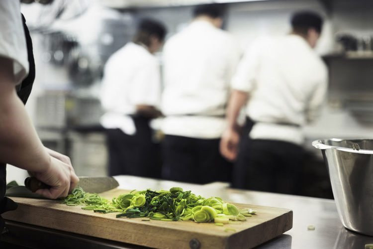Cropped image of female chef chopping leek on cutting board with colleagues in background at restaurant