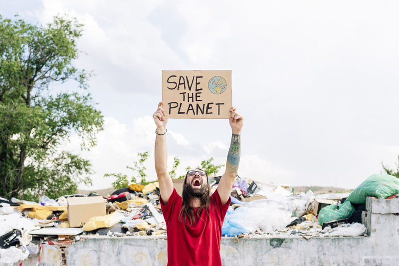 Hippie man shouting while holding save the planet cardboard near garbage dump