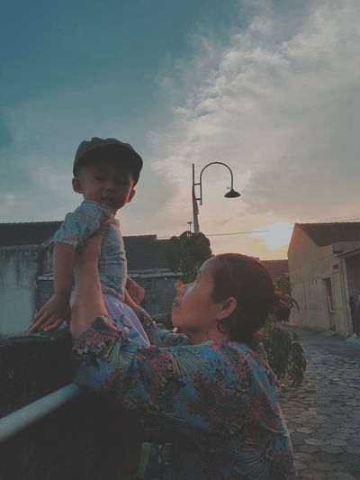 Side view of mother holding cute son sitting on retaining wall against cloudy sky during sunset