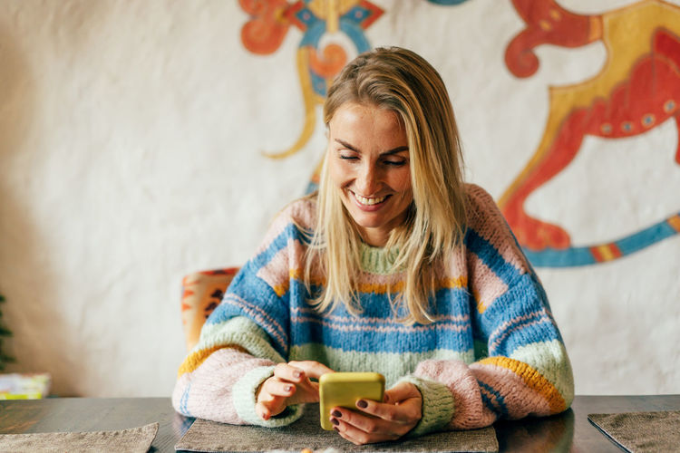A young woman with a mobile phone at a table in a cafe laughs and watches social media.