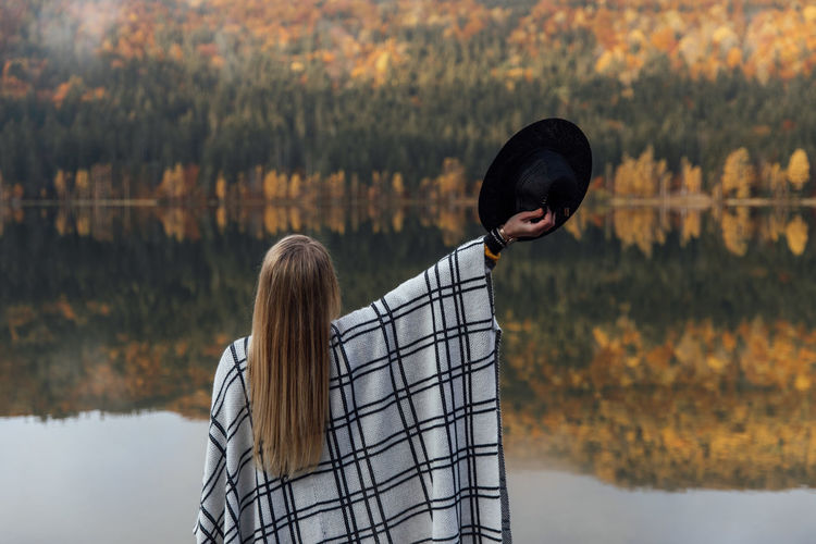 Blonde female wearing poncho and hat,standing with raised arm on background of autumn forest lake.