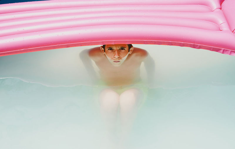 Boy in a pool taking a bath under a pink flat looking at camera.