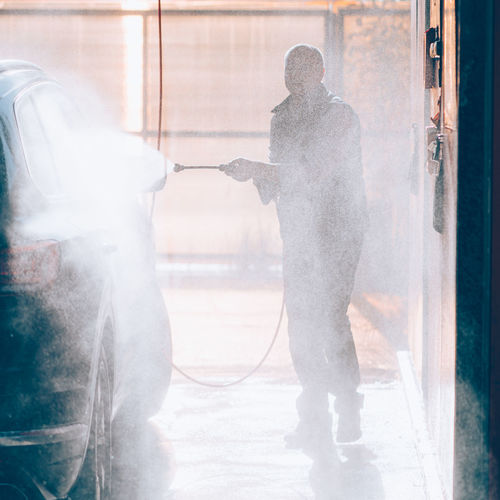 Man washes car at car wash, pours water from hose. sunlight and water splashes in contour