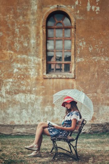 Side view full length of young woman with umbrella reading book while sitting against building on bench