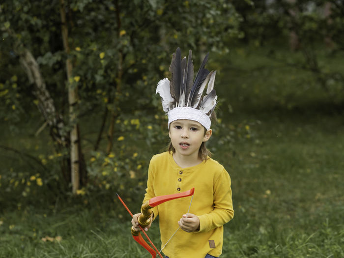 Little boy is playing american indian on field. kid has headdress made of feathers,bow with arrows. 