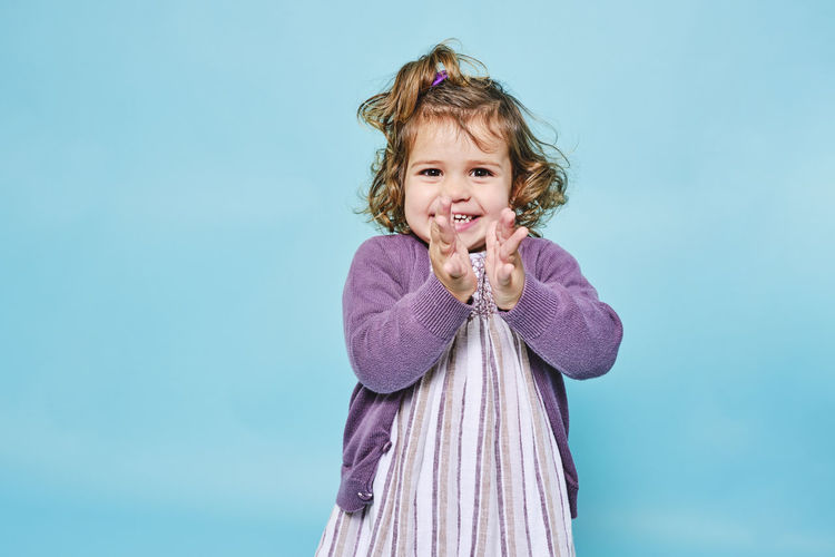 Cheerful small child in purple dress and knitted cardigan smiling at camera and clapping while standing alone against light blue background in modern studio