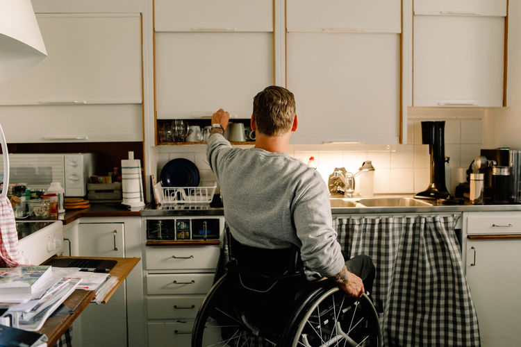 Rear view of disabled man opening cabinet in kitchen at home