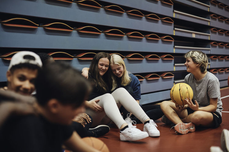 Smiling teenage girl sitting with cross-legged looking at teenage female friends embracing each other in basketball cour