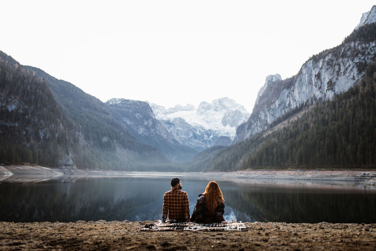 Back view of unrecognizable couple in warm clothes sitting on shore of peaceful lake and enjoying spectacular landscape of snowy mountains in autumn morning